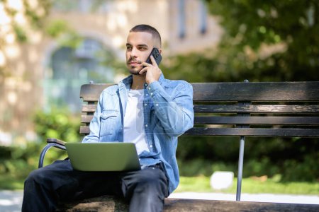 Foto de Hombre joven que utiliza el ordenador portátil para estudiar o trabajar al aire libre. Joven hombre vistiendo ropa casual y sentado en el banco en el parque público y teniendo una llamada. - Imagen libre de derechos