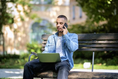 Téléchargez les photos : Jeune homme utilisant un ordinateur portable pour étudier ou travailler à l'extérieur. Jeune homme portant des vêtements décontractés et assis sur le banc dans le parc public et ayant un appel. - en image libre de droit