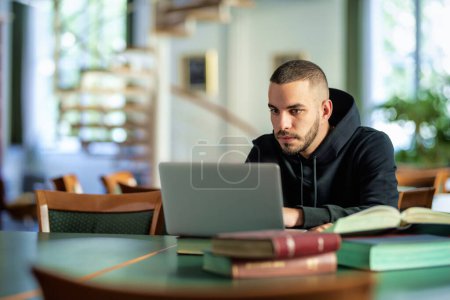 Téléchargez les photos : Jeune homme confiant assis dans la bibliothèque universitaire et apprenant. Étudiant caucasien utilisant un ordinateur portable et des livres. - en image libre de droit
