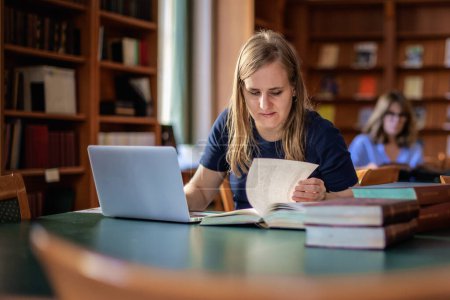 Téléchargez les photos : Une jeune femme malvoyante assise et étudiant à la bibliothèque universitaire. Jeune femme utilisant des livres et un ordinateur portable. - en image libre de droit