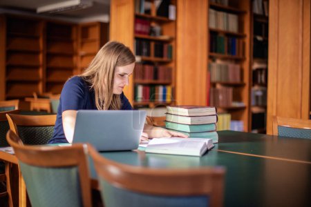 Téléchargez les photos : Une jeune femme malvoyante assise et étudiant à la bibliothèque universitaire. Jeune femme utilisant des livres et un ordinateur portable. - en image libre de droit