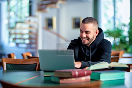Foto de Joven confiado sentado en la biblioteca universitaria y aprendiendo. Estudiante caucásico usando laptop y libros. - Imagen libre de derechos