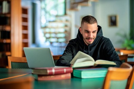 Photo for Confident young man sitting in the university library and learning. Caucasian student using laptop and books. - Royalty Free Image