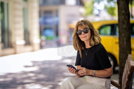 Téléchargez les photos : Attrayant femme aux cheveux blonds est assis sur un banc dans la rue et en utilisant un téléphone mobile. Joyeux sourire féminin portant des lunettes de soleil et des messages texte. - en image libre de droit