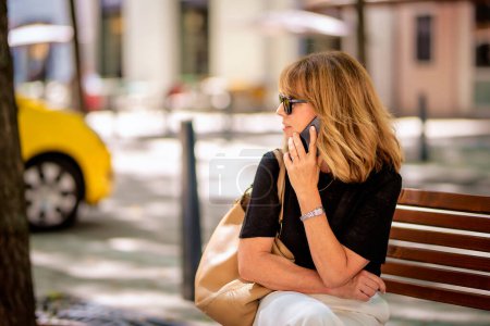 Téléchargez les photos : Attrayant femme aux cheveux blonds est assis sur un banc dans la rue et en utilisant un téléphone mobile. Joyeux sourire féminin portant des lunettes de soleil et passer un appel. - en image libre de droit