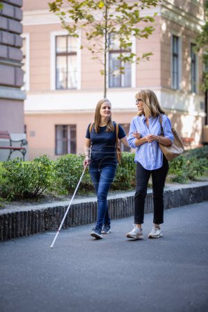 Foto de Mujer con discapacidad visual caminando con una amiga en la calle de la ciudad. Mujer caucásica madura ayudando a su amigo que es discapacitado visualmente. Longitud completa. - Imagen libre de derechos