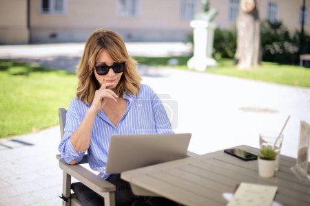 Photo for A middle-aged blond haired woman sitting on the terrace of a cafe and using a laptop for work. Confident female looking thoughtfully. - Royalty Free Image