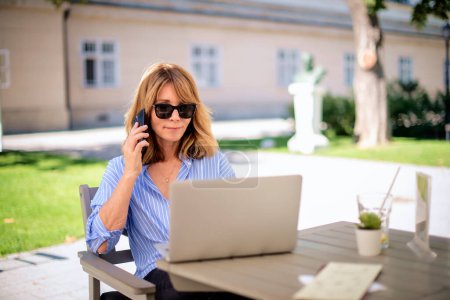 Foto de Una rubia de mediana edad sentada en la terraza de un café y usando un portátil. Sonriente hembra usando laptop y celular para el trabajo. - Imagen libre de derechos