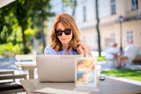 Photo for A middle-aged blond haired woman sitting on the terrace of a cafe and using a laptop. Cheerful smiling female using laptop and cellphone for work. - Royalty Free Image