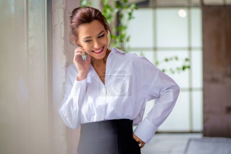 Photo for Portrait of a brown haired middle-aged woman wearing a white shirt and black pants and standing by the window. Attractive female cheerful smiling. - Royalty Free Image