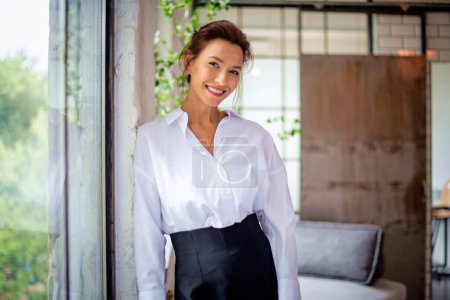 Photo for Portrait of a brown haired middle-aged woman wearing a white shirt and black pants and standing by the window. Attractive female looking at camera and cheerful smiling. - Royalty Free Image