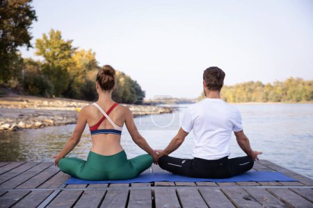 Téléchargez les photos : Vue arrière d'une femme et d'un homme assis en position lotus sur un tapis de yoga extérieur. Toute la longueur d'un couple méditant sur la jetée au bord de la rivière. - en image libre de droit