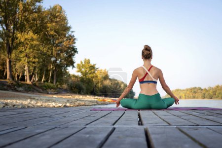 Téléchargez les photos : Vue arrière d'une femme assise en position lotus sur un tapis de yoga extérieur. Toute la longueur d'un entraîneur de yoga féminin méditant sur la jetée au bord de la rivière. - en image libre de droit