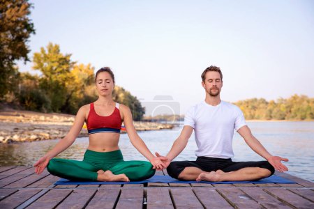 Photo for Shot of a woman and a man sitting in lotus position on yoga mat outdoor. Full length of a couple meditatiing on the pier on riverside. - Royalty Free Image