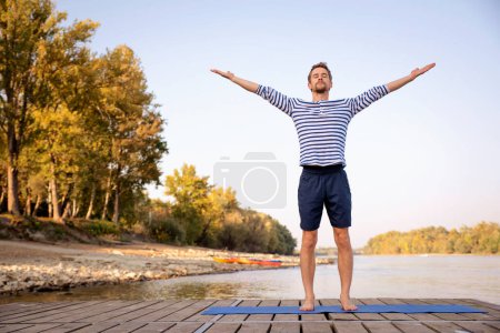 Foto de Hombre de mediana edad practicando yoga al aire libre. Hombre caucásico usando esterilla de yoga y de pie con los brazos levantados en la naturaleza. Saludo al sol. - Imagen libre de derechos