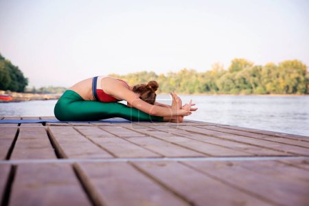 Téléchargez les photos : Toute la longueur d'une femme pratiquant le yoga sur la jetée au bord de la rivière. Dame athlétique étirement sur tapis de yoga. - en image libre de droit