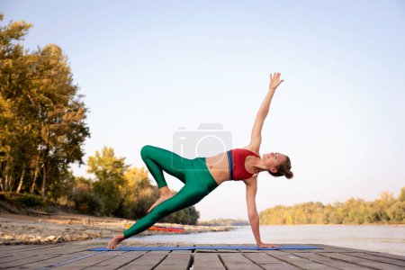 Téléchargez les photos : Toute la longueur d'une femme pratiquant le yoga sur la jetée au bord de la rivière. Dame athlétique étirement et échauffement sur tapis de yoga, - en image libre de droit