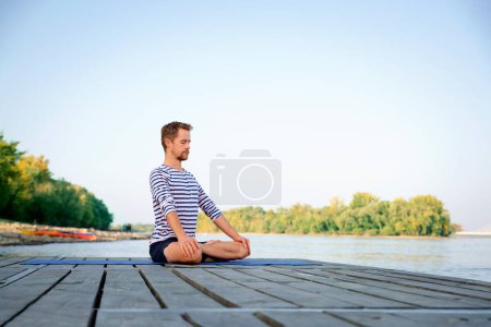 Foto de Longitud completa del hombre caucásico haciendo yoga en el muelle junto al lago. Hombre maduro practicando la posición de loto y relajándose. - Imagen libre de derechos