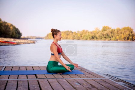 Téléchargez les photos : Femme aux cheveux blonds médite assis en position lotus sur tapis de yoga en plein air. Longueur totale d'un entraîneur de yoga féminin pratiquant le yoga sur la jetée sur le bord de la rivière. - en image libre de droit