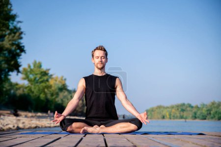 Foto de Longitud completa del hombre caucásico practicando yoga en el muelle junto al lago. Hombre maduro practicando la posición de loto en embarcadero contra el cielo azul. - Imagen libre de derechos