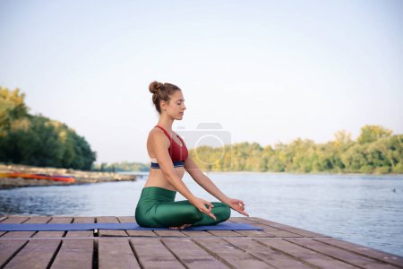 Téléchargez les photos : Femme aux cheveux blonds médite assis en position lotus sur tapis de yoga en plein air. Longueur totale d'un entraîneur de yoga féminin pratiquant le yoga sur la jetée sur le bord de la rivière. - en image libre de droit