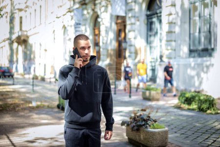 Photo for Cropped shot of a handsome young man using his cellphone while walking on the street in the city. Confident male wearing hoodie and making a call. - Royalty Free Image