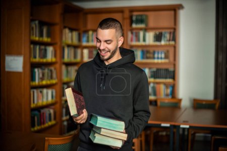 Foto de Joven parado en la biblioteca de la universidad y sosteniendo libros en su mano. Hombre guapo con sudadera con capucha. - Imagen libre de derechos