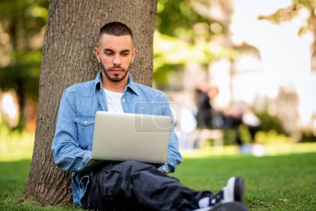 Téléchargez les photos : Jeune homme utilisant un ordinateur portable pour étudier ou travailler en plein air. Jeune homme portant des vêtements décontractés et assis sur l'herbe dans le parc public. Plan complet. - en image libre de droit