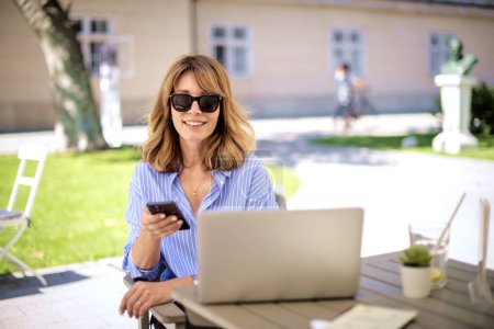 Foto de Mujer de negocios segura sentada en la cafetería al aire libre y trabajando. Atractiva hembra usando laptop y mensajería de texto. - Imagen libre de derechos