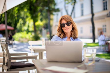 Photo for A middle-aged blond haired woman sitting on the terrace of a cafe and using a laptop. Confident female wearing sunglasses and blue shirt. - Royalty Free Image