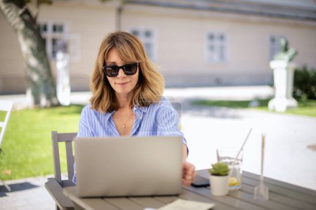 Foto de Una rubia de mediana edad sentada en la terraza de un café y usando un portátil. Mujer segura con gafas de sol y camisa azul. - Imagen libre de derechos