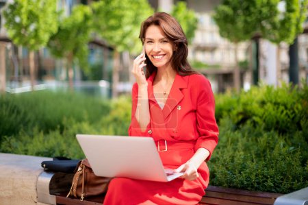 Téléchargez les photos : Femme aux cheveux bruns assise sur un banc en ville et utilisant un ordinateur portable et un smartphone pour le travail. Heureuse femme d'âge moyen portant des vêtements rouges et ayant un appel. - en image libre de droit