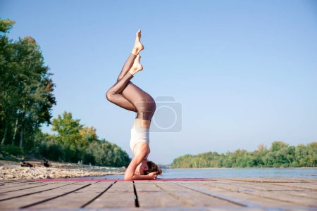 Téléchargez les photos : Jeune femme faisant tête haute surplombant la rivière. Longueur totale d'un entraîneur de yoga féminin pratiquant le yoga sur la jetée sur le bord de la rivière. - en image libre de droit