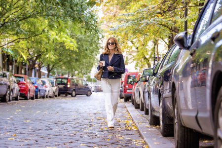 Photo for Full length of an attractive middle aged woman holding paper coffee cup  walking  on street - Royalty Free Image