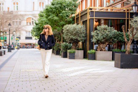 Photo for A middle-aged woman with blonde hair is walking down the street in the city centre. Attractive female wearing sunglasses and blue coat. Full length shot. - Royalty Free Image