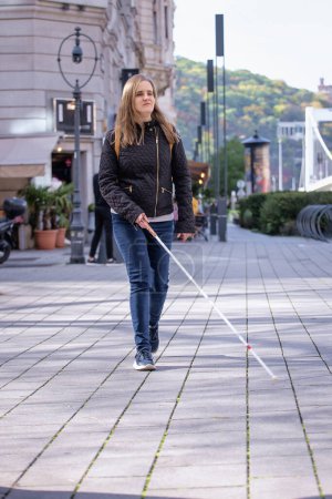 Foto de Retrato de mujer ciega con bastón blanco caminando por la calle. Una mujer con discapacidad visual que viste ropa casual y usa su bastón para caminar por la calle. Disparo completo. - Imagen libre de derechos