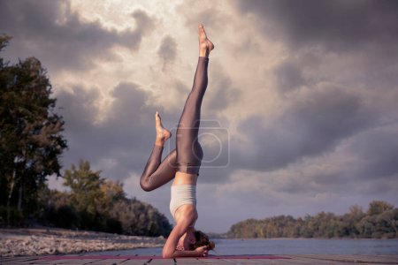 Téléchargez les photos : Jeune femme en position debout surplombant la rivière. Longueur totale d'un entraîneur de yoga féminin pratiquant le yoga sur la jetée sur le bord de la rivière. - en image libre de droit
