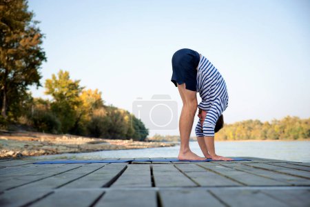 Foto de Hombre de mediana edad practicando yoga al aire libre. Hombre atlético estirándose en el muelle junto al río. - Imagen libre de derechos