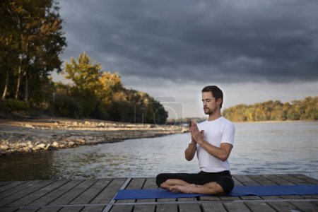 Téléchargez les photos : Toute la longueur de l'homme caucasien faisant du yoga sur jetée par lac. Homme mûr pratiquant la position du lotus et se détendre. - en image libre de droit