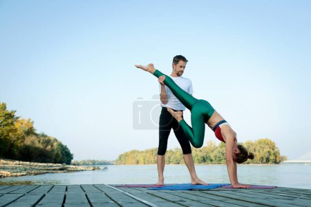 Foto de Una foto de una mujer y un hombre haciendo yoga acrobático al aire libre. Longitud completa de una pareja practicando yoga en el muelle a orillas del río. - Imagen libre de derechos