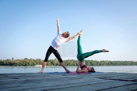 Foto de Una foto de una mujer y un hombre haciendo yoga acrobático al aire libre. Longitud completa de una pareja practicando yoga en el muelle a orillas del río. - Imagen libre de derechos