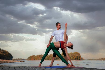 Photo for Shot of a woman and a man doing acrobatic yoga outdoor. Full length of a couple practicing yoga on the pier on riverside. - Royalty Free Image