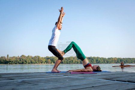 Photo for Shot of a woman and a man doing acrobatic yoga outdoor. Full length of a couple practicing yoga on the pier on riverside. - Royalty Free Image
