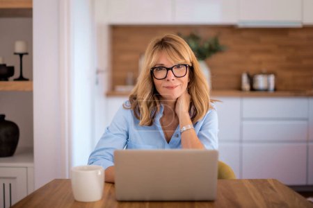 Blonde Frau sitzt zu Hause und benutzt Laptop für die Arbeit. Home office. Selbstbewusste Geschäftsfrau mit Webkonferenz oder Surfen im Internet. 