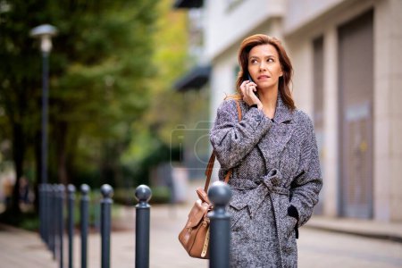 Téléchargez les photos : Femme utilisant smartphone et marcher à l'extérieur dans la rue de la ville le jour de l'automne. Jolie femelle portant un manteau en tweed et ayant un appel. - en image libre de droit