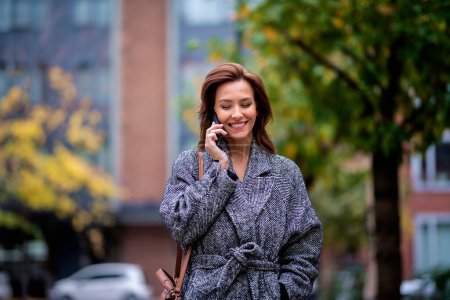 Foto de Mujer usando smartphone y caminando al aire libre en la calle de la ciudad en el día de otoño. Atractiva hembra usando abrigo de tweed y teniendo una llamada. - Imagen libre de derechos