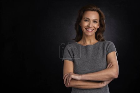 Téléchargez les photos : Portrait studio d'une femme aux cheveux bruns portant une robe grise et souriant sur fond sombre isolé. Espace de copie. - en image libre de droit
