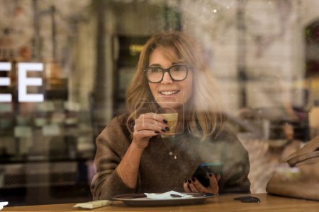 Photo for Smiling blond haired woman sitting in cafe and using her smartphone. Attractive female drinking coffee and photographed through the window. - Royalty Free Image