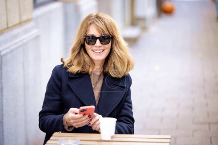 Foto de Mujer sentada en una mesa frente a un café y usando su teléfono inteligente mientras bebe una taza de café. Mujer segura usando ropa casual y gafas de sol. - Imagen libre de derechos
