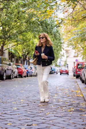 Foto de Longitud completa de una atractiva mujer de mediana edad sosteniendo una taza de café de papel y su teléfono móvil en la mano y caminando por la calle de la ciudad. - Imagen libre de derechos